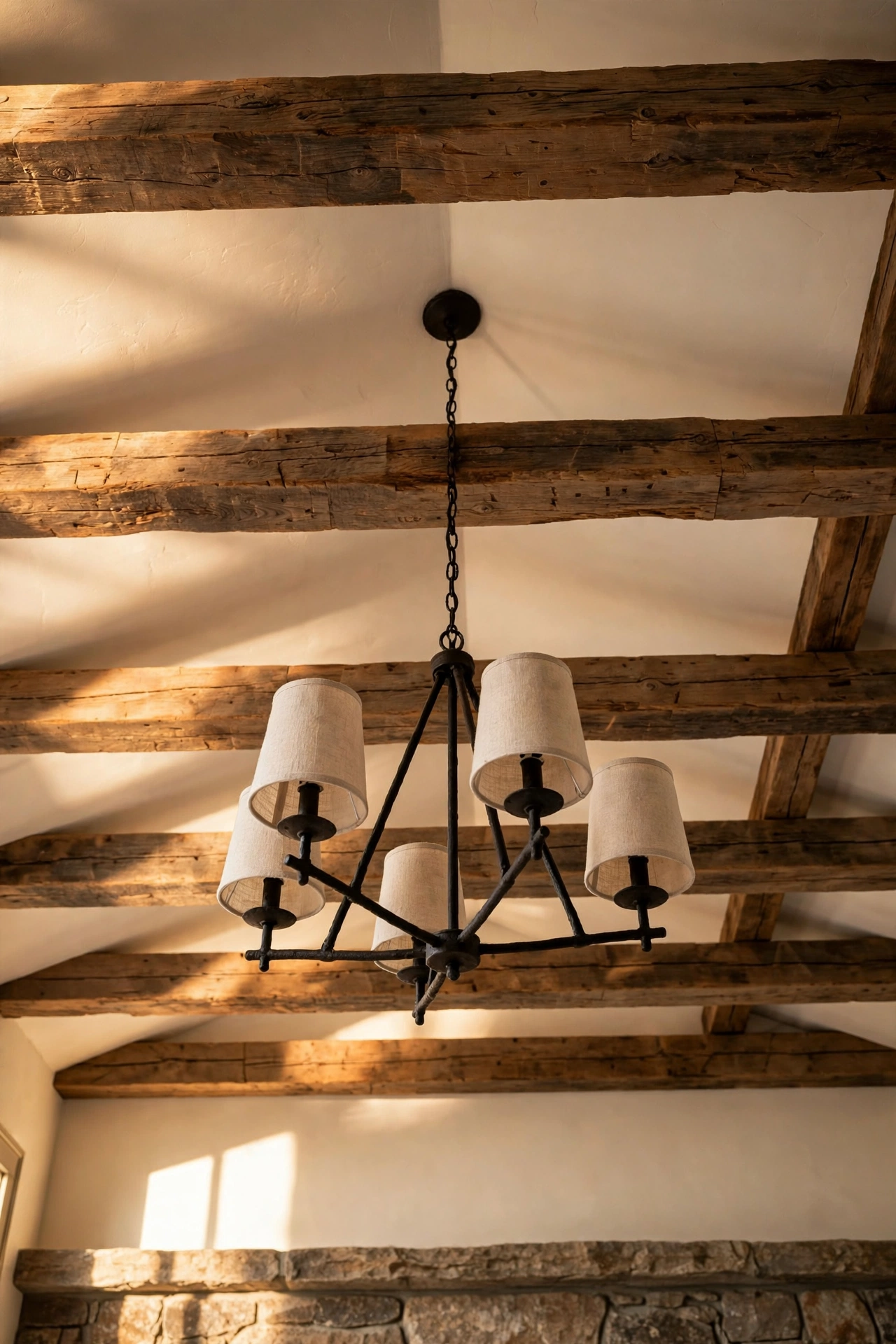 Reclaimed wood beams in walnut across a warm white ceiling with an iron chandelier and linen shades above a stone fireplace